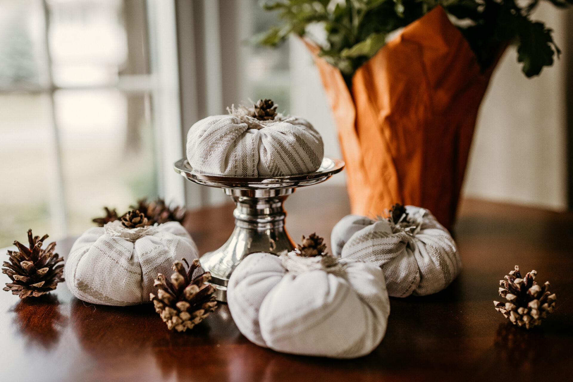 A set of 4 linen pumpkins with neutral color stripes topped with mini pinecones are photographed in front of a window.