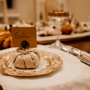 An neutral color linen pumpkin is placed on a gold rimmed plate that is part of an elegant table setting. The pumpkin is topped with a metal swirl that is holding a Kraft paper place card bearing the name Sofia.