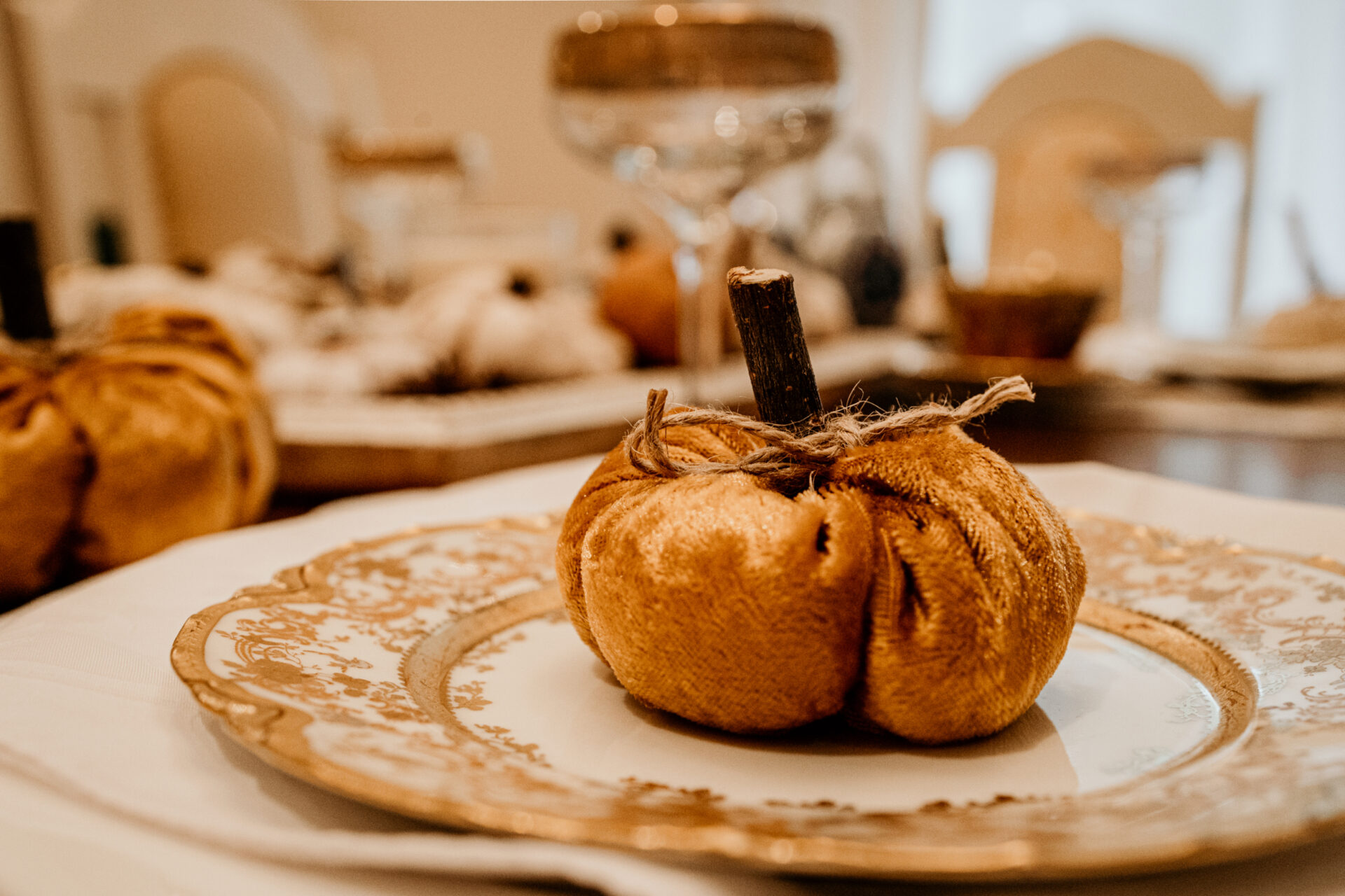 An orange velvet pumpkin with a wooden stick as its stem is placed on a gold rimmed plate that is part of an elegant table setting.