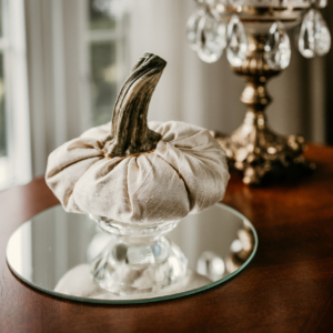 A silk pumpkin with a real stem is propped on a crystal stand sitting on a round mirror and photographed in front of a window.
