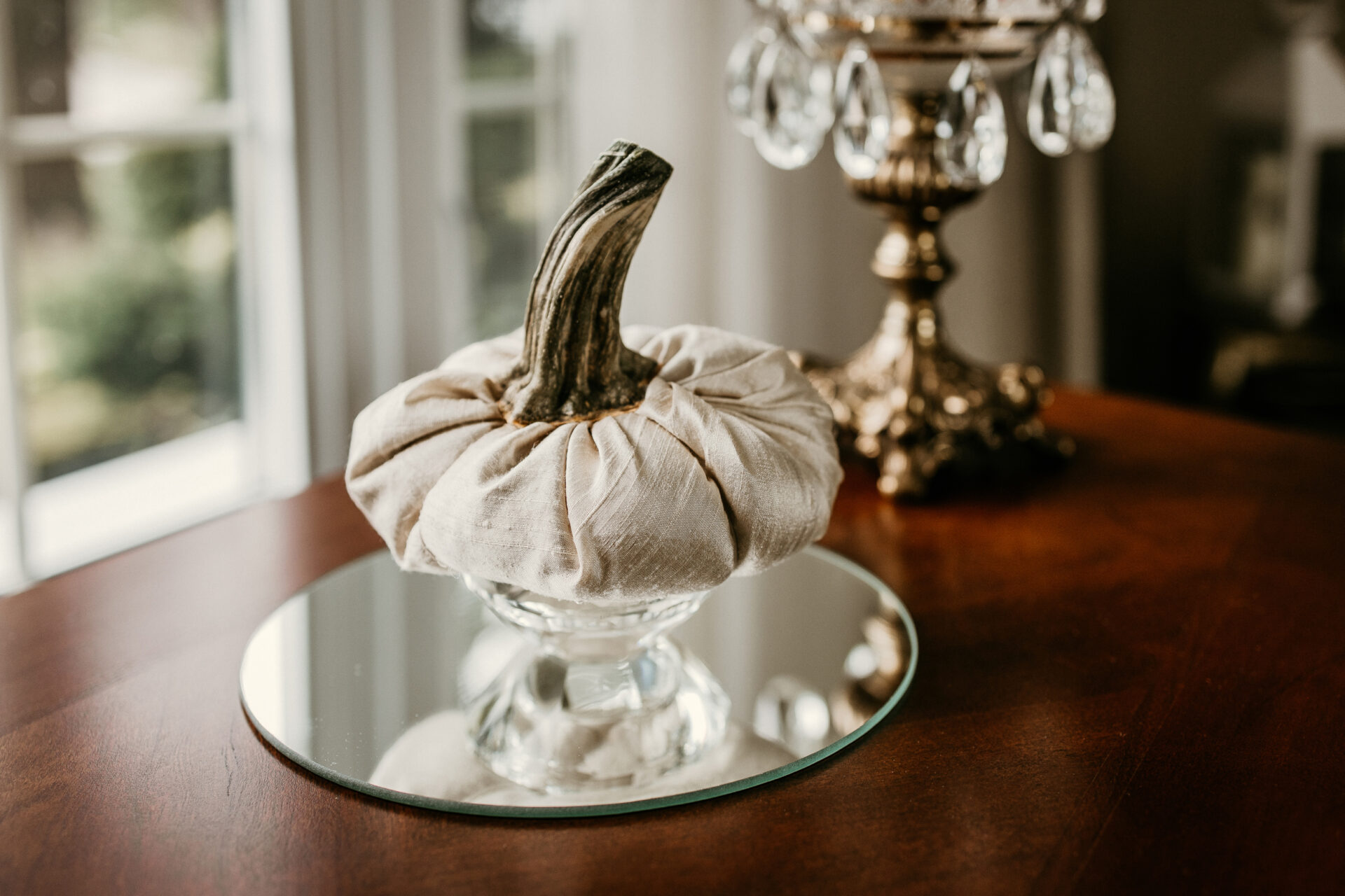 A silk pumpkin with a real stem is propped on a crystal stand sitting on a round mirror and photographed in front of a window.