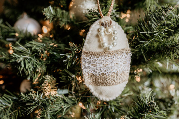 An oval shaped felt ornament decorated with pearls, lace and burlap hanging on a Christmas tree