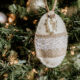 An oval shaped felt ornament decorated with pearls, lace and burlap hanging on a Christmas tree