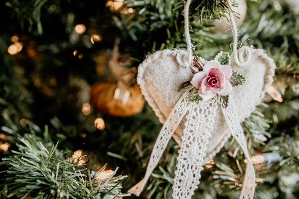 A heart shaped felt ornament decorated with a porcelain rose and lace ribbon is hanging on a Christmas tree