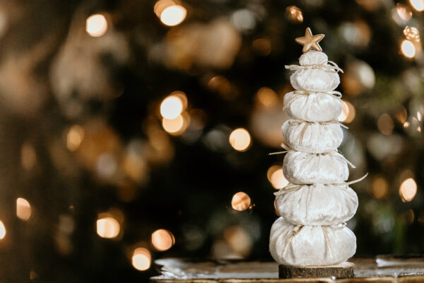 An off white velvet tree with a gold star on top sitting on a real wood base is pictured in front of a Christmas tree