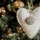 A heart shaped felt ornament decorated with an antique broach and lace ribbon is hanging on a Christmas tree