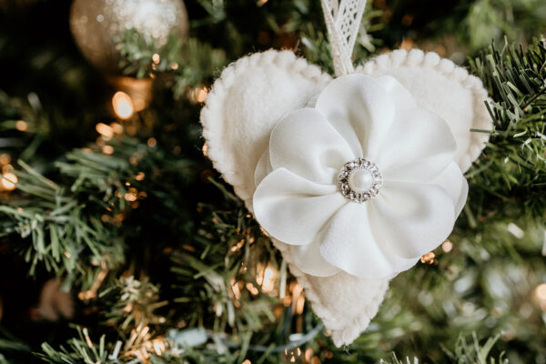 A heart shaped felt ornament decorated with a fabric flower and jeweled center is hanging on a Christmas tree